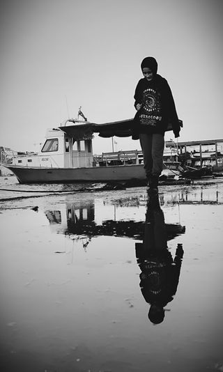 Black and white photo of a person walking near a boat, with their reflection visible in a puddle. 
