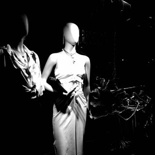Black and white image of two mannequins in a dark window display. One wears a knotted shirt and necklace, the other a sleeveless dress. Dramatic shadows create a moody ambiance.