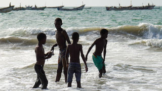 Four young boys playing in the ocean water with boats on background.