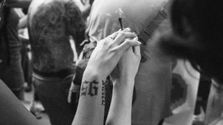 Black and white photo of tattooed arms holding a cigarette in a crowd. Intimate, relaxed vibe with tattoo art detail and faint smoke.