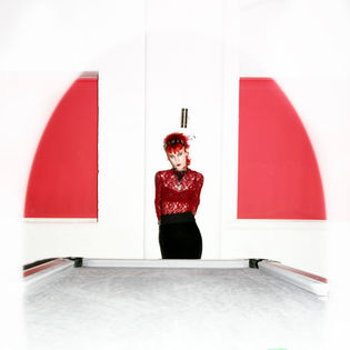 A person with red hair wearing a red lace top and black skirt stands against a white and red wall, with a pool table in the foreground.