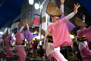 Dancers in pink kimonos and straw hats perform joyfully in a lively street festival at night, with onlookers and bright lights in the background.