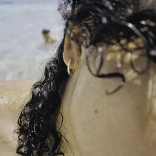 Close-up of a person with wet, curly hair and a small hoop earring, standing in shallow ocean water. A blurred figure swims in the background, suggesting a beach setting.