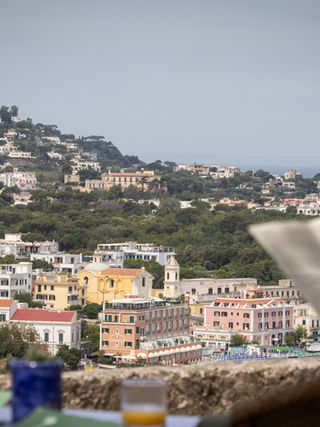 Man reads newspaper, overlooking Capri town, sunny day, glasses, hat.