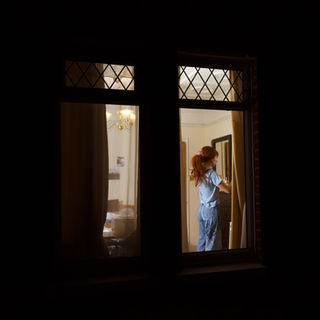 A person with red hair and a ponytail stands inside a warmly lit room, viewed through a window, closing the curtains. The scene is cozy and intimate.