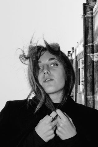 A woman with windblown hair stands against a building, looking thoughtfully off to the side. She wears a dark coat and holds her hair while gazing ahead. The image is in black and white.