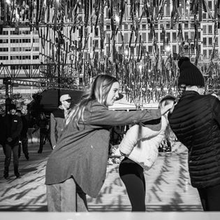 Black and white image of people ice skating under hanging lights. They appear joyful and playful against a backdrop of a city building.