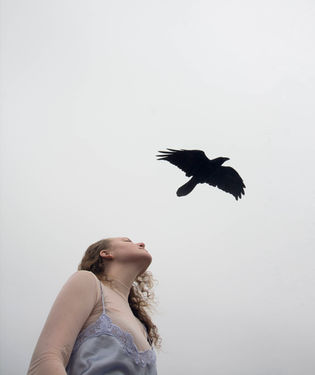 A woman gazes upward as a black bird flies overhead against a pale sky. Her expression is serene, conveying a sense of freedom and introspection.