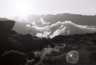 Black and white image of a serene mountain landscape; clouds drift above jagged peaks at sunset, with a small stream flowing in the foreground.