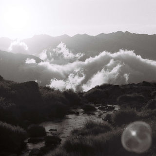Black and white image of a serene mountain landscape; clouds drift above jagged peaks at sunset, with a small stream flowing in the foreground.