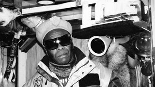Black and white photo of a man in sunglasses and a beret posing in an eclectic room. He wears a jacket with a patch. 