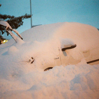A car buried under heavy snow at dusk, with only its side mirror, wipers, and outline visible. Streetlights glow softly in the background, evoking a chilly, serene winter scene.