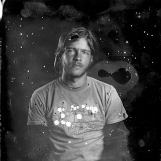 Black and white portrait of a man with long hair, wearing a graphic T-shirt. They sit against a textured, speckled backdrop. 