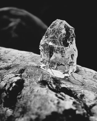 A clear, jagged crystal or mineral sits on a rough rock surface, photographed in black and white.