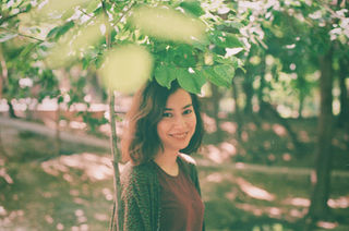 A woman with brown hair smiles under a leafy tree in a sunlit park. Dappled light filters through the leaves, creating a serene and cheerful atmosphere.