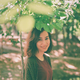 A woman with brown hair smiles under a leafy tree in a sunlit park. Dappled light filters through the leaves, creating a serene and cheerful atmosphere.