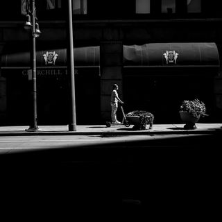 Black and white photo of a man on a scooter in sunlight, beside large planters, in front of a dark bar entryway, creating strong contrasts.