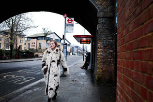A woman in a long, spotted fur coat walks on a city sidewalk under a bridge. A bus stop and another person are visible in the background. Overcast day.