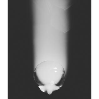 A single, glossy water droplet in grayscale, reflecting light, is centered against a blurred, dark background. The mood is calm and reflective.