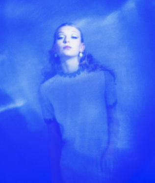 A woman stands in an ethereal blue haze, wearing a patterned top. Her expression is serene, evoking a dreamy, mystical ambiance.