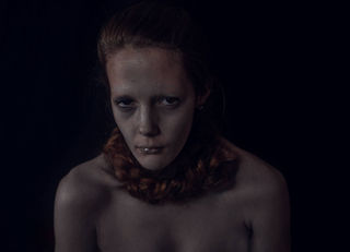 A somber woman with pale skin and dark circles under her eyes stares forward against a black background. Her expression is intense and haunting.