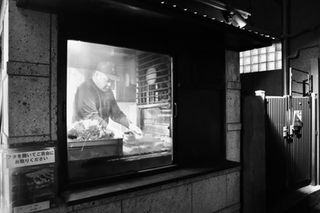 Black and white photo of a man cooking inside a small, lit window, creating a cozy, intimate street scene. 