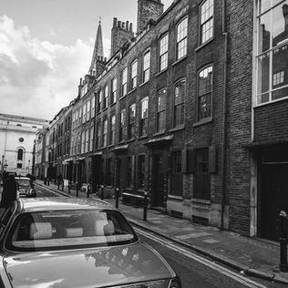 Black and white photo of a deserted city street lined with historic brick buildings and a distant church spire. Cars parked along the curb on a cloudy day.