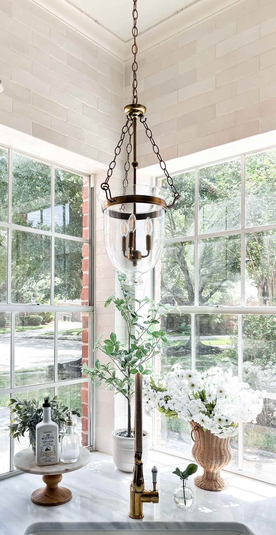 corner kitchen sink with tall windows, lantern style brass light fixture, marble countertops, soap and lotion styled on top of cake stand, wicker vase with spring flowers, and faux tree