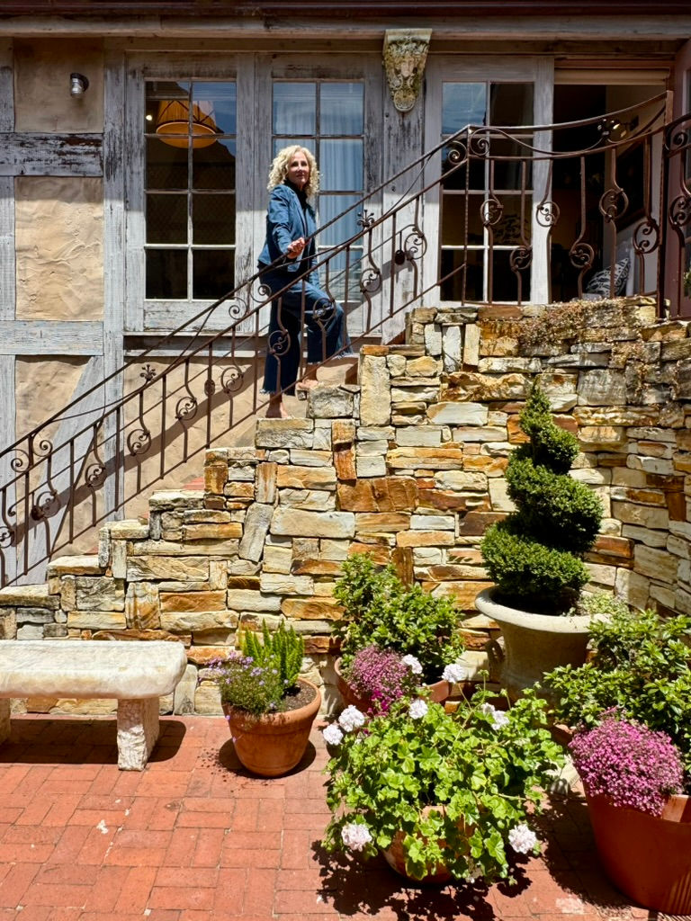 Woman in blue climbs a stone staircase outside a rustic building. Potted plants and flowers on a brick patio; bright, sunny ambiance.
