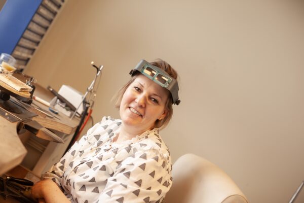 Kvinne smiler med forstørrelsesbriller. Brenda Meinen, smykke designer, ved benken.