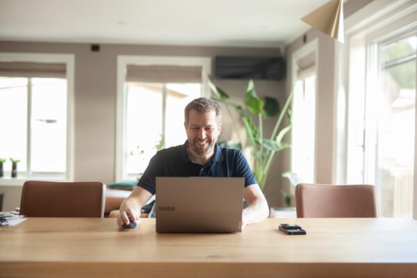 Smilende mann jobber med bærbar PC ved et bord i et hjem.