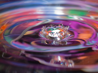 a drop going into water with concentric circles coming out. The water has a rainbow color quality to it, vibrant pinks and greens and oranges and blues and blacks. 