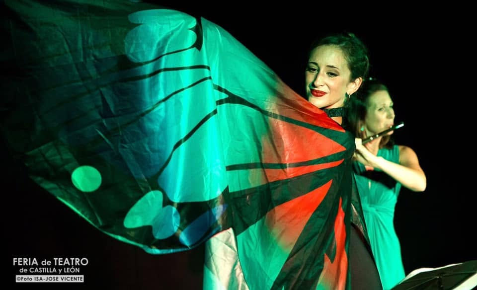 Two women perform on a dark stage; one waves green-red-black fabric while another plays flute. Feria de Teatro de Castilla y León.