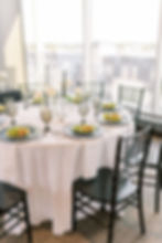 Elegant table setting with black chairs, white tablecloth, green salads on black plates, and tall candles in a bright room with large windows.