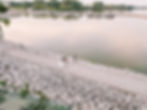 Bride and groom dip on a lakeside path surrounded by rocks. Boats docked in calm water, with lush greenery in the background. Romantic mood.