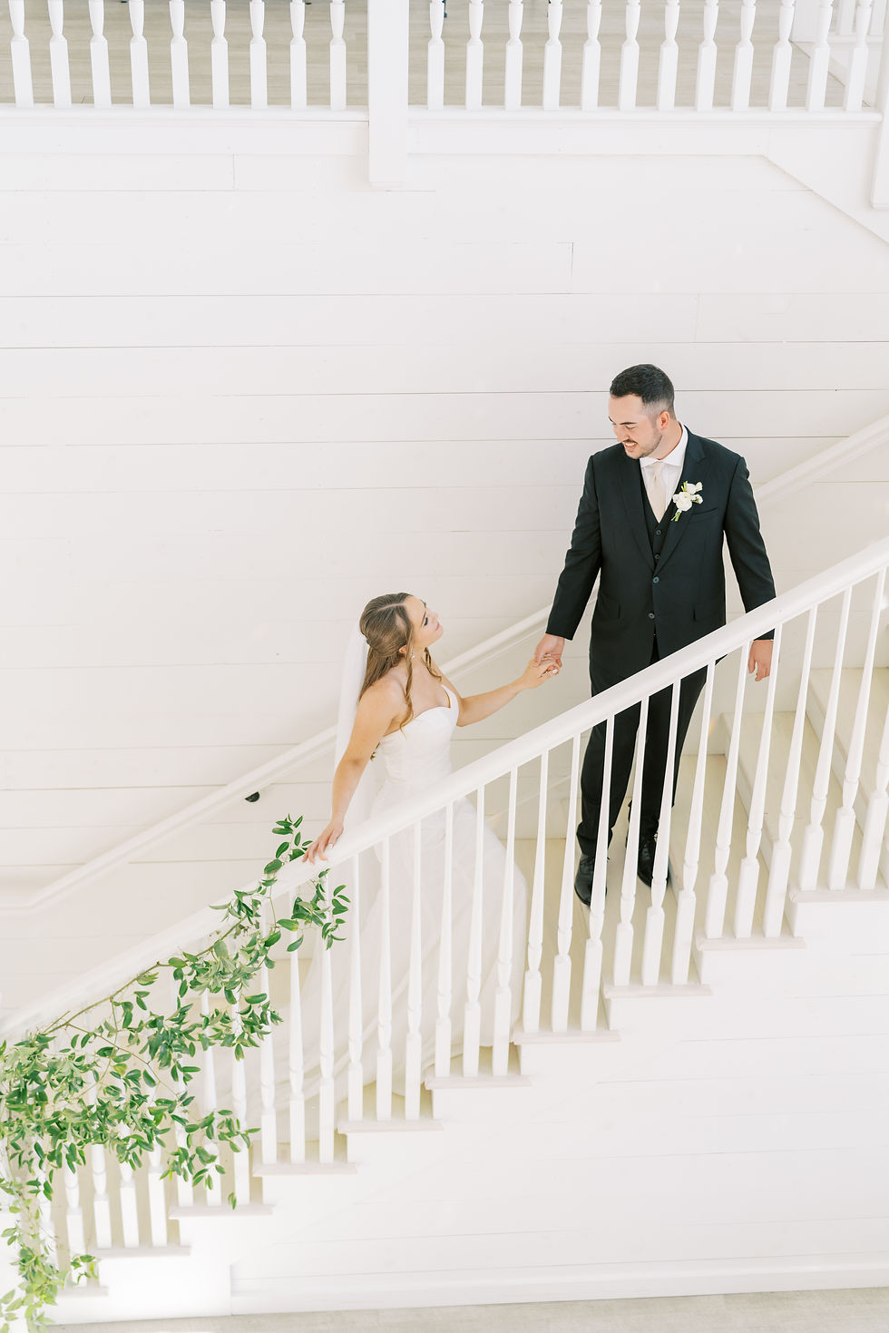 Bride and groom hold hands on white staircase adorned with greenery. The bride smiles, wearing a white dress, in a bright, joyful setting.