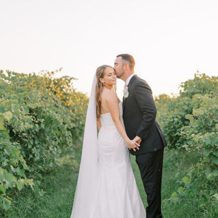 A groom kissing his bride on the cheek and holding hands in the middle of a vineyard.