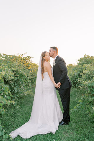 A groom kissing his bride on the cheek and holding hands in the middle of a vineyard.