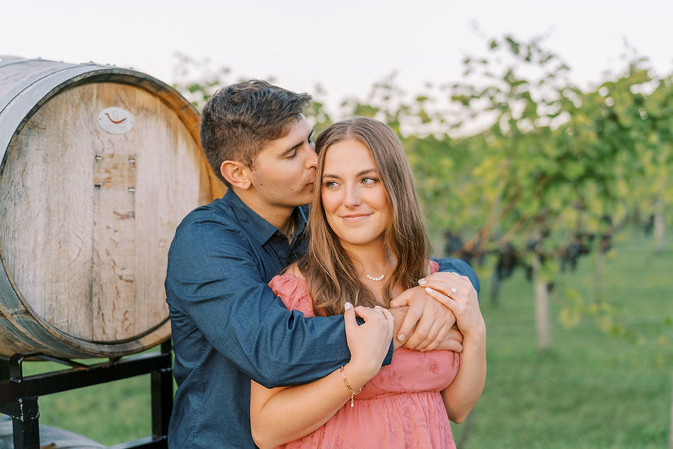 A couple embraces by a vineyard barrel. The man kisses the woman's cheek, both wearing casual outfits. Lush vineyard in the background.