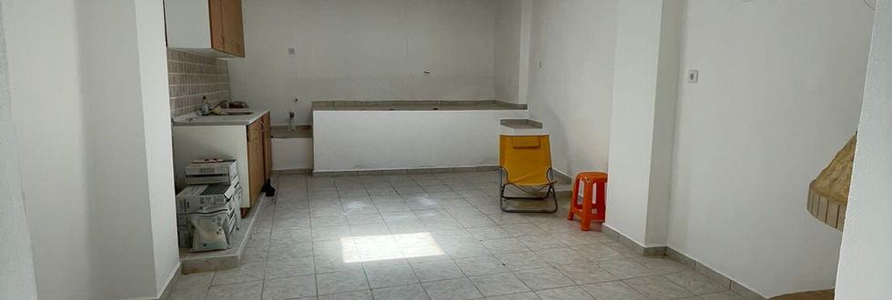 Open-concept setting in a stone-built house for sale in a traditional village in Crete, capturing the small kitchen and living room with a fireplace. The view is from the entrance of the house, revealing a cozy and welcoming living space within this charming property.