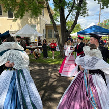 Three couples from the Dolina Polish Folk Dance Group perform a traditional waltz outside on a shaded area of pavement. The women wear elaborate lace-trimmed blouses and long, flowing skirts in rich colors—blue, purple, and pink—paired with white aprons and ribbon sashes. The men are dressed in black coats and hats accented with colored sashes. A crowd of festival-goers watches joyfully in the background under red, white, and blue tents on a sunny afternoon.