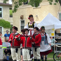 A group of Dolina Polish Folk Dancers in traditional costumes performing a lift during a cultural dance. Three men in red embroidered jackets and white pants support a smiling female dancer seated on their shoulders. She wears a black and yellow bodice, a long patterned skirt, and an elaborate red and black headpiece with matching beads. Two other women in traditional dress stand behind them, smiling, with a “Take A Chance” raffle booth visible in the background.
