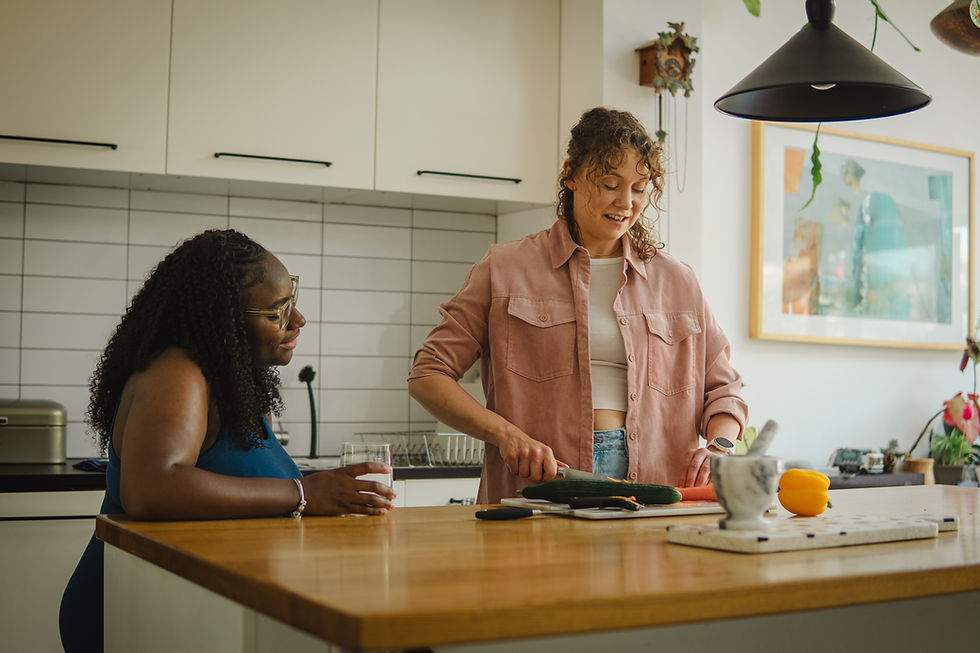 Two women in a bright kitchen, one slicing cucumber while the other watches with a glass; warm, relaxed mood
