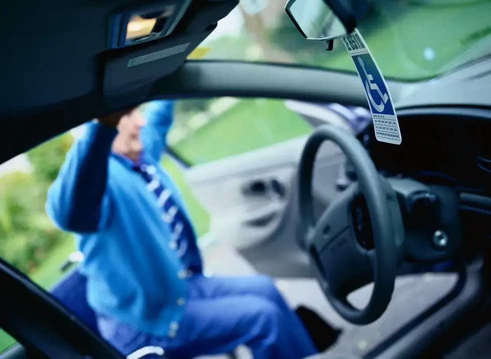 A person in a blue outfit, seated in a wheelchair, transfers into a car with a hanging disabled permit. The background is green and blurred.