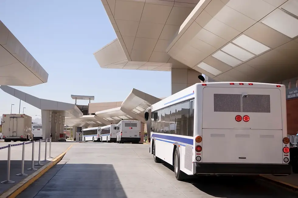 White buses lined up under a modern, angular roof at a bus terminal. Sunlit day, clear sky, and visible sign: "Unloading Area."