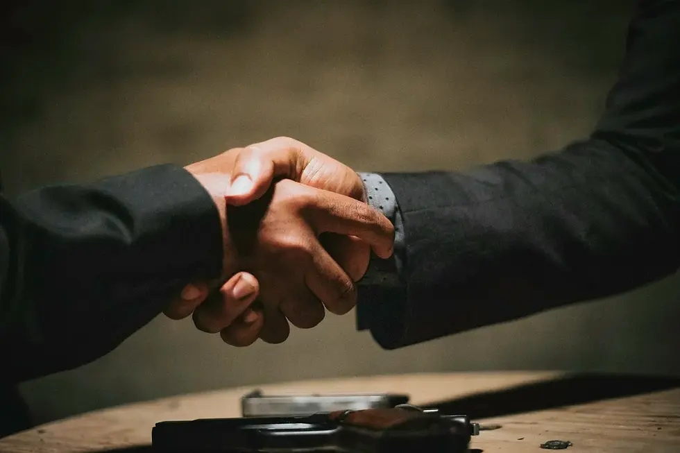 Two individuals in dark suits shake hands over a table with a handgun and a blurred background, creating a tense and serious mood.