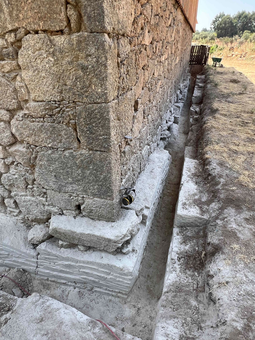 Long, dug-out drainage channel beside a granite stone structure showing exposed footings and rubble.