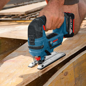 Close-up of a Bosch GST 18 V-Li B cordless jigsaw cutting through a wooden plank while a person holds the board steady.