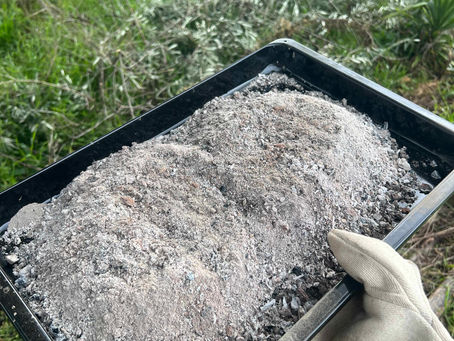 Black La Nordica tray with fine grey ash being carried outdoors over a grassy patch.