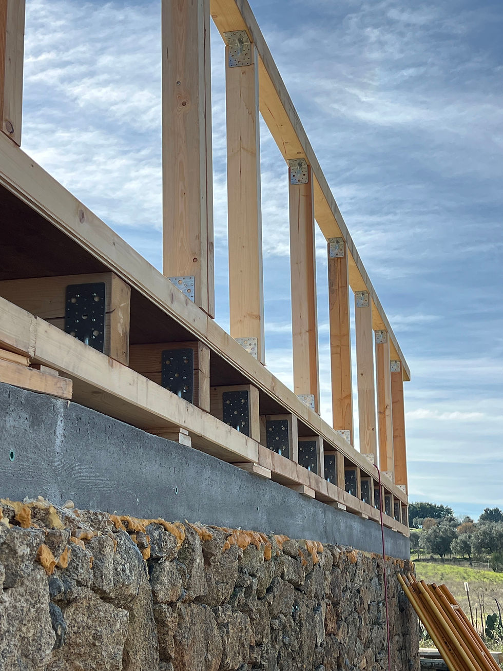 Timber stud wall fixed to a concrete bond beam above a stone foundation, blue sky behind.